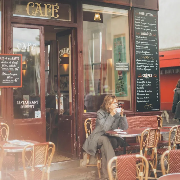 A middle aged women sitting at a French cafe drinking coffee and people watching
